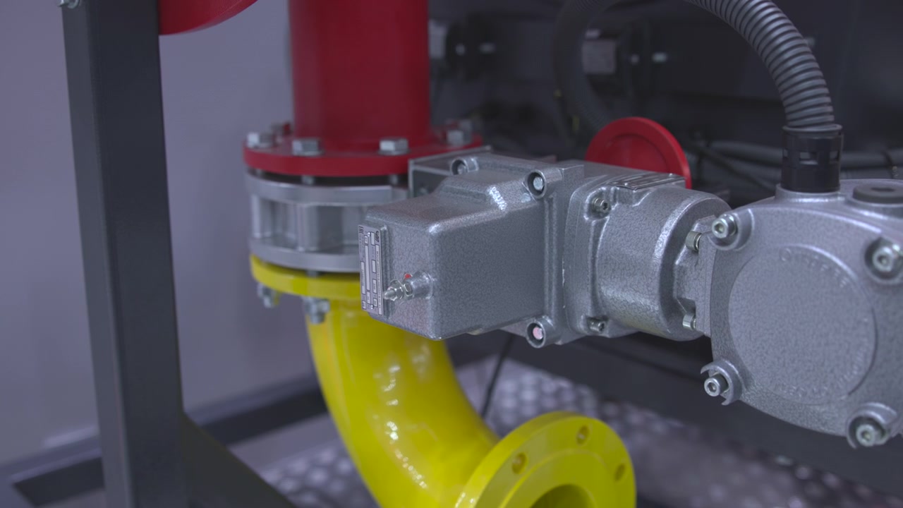Gros plan sur un réducteur industriel et un carter de moteur raccordés rigidement à un ensemble de tuyauterie à brides à l'intérieur d'un local technique, avec des boulons, un conduit, un tuyau jaune, un raccord rouge et une surface métallique usée