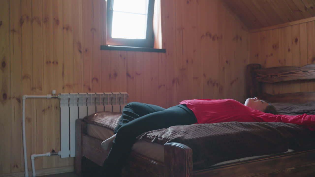 young lady in red shirt enters wooden room and gently walks toward bed before sitting down in calm and, relaxed motion, surrounded by warm rustic tones and soft indoor lighting