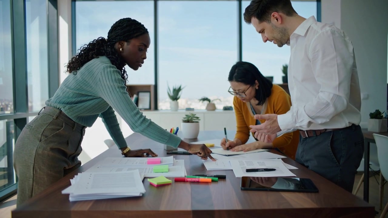 Pointing trio in business wear sorting papers at office table with tablet and notes finalizing plan