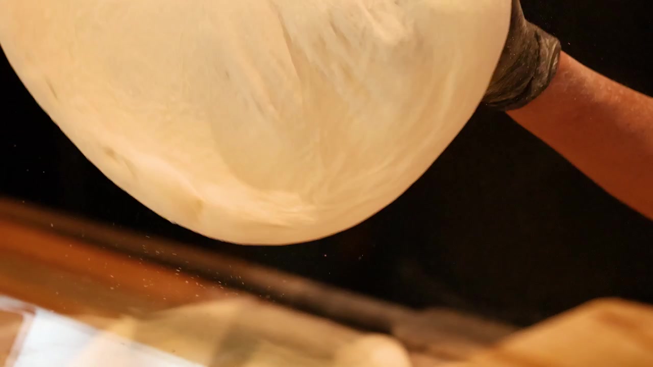 Chef wearing black gloves stretches and tosses pizza dough in a dimly lit professional kitchen.
