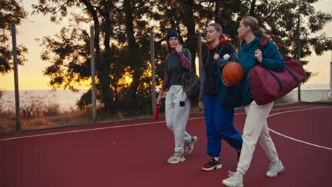 side view of a happy blonde girl in a turquoise hoodie walking and carrying a basketball while chatting with her two friends after a basketball match in the evening at sunset