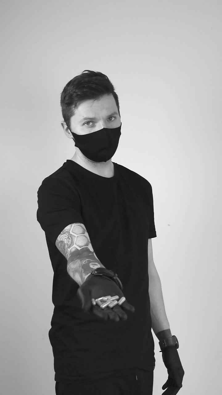 Grayscale video of white man with health protective face mask and black gloves shoving a bunch of medicine pills or capsules on his hand and then throws them into the air in slow motion, pills flying.