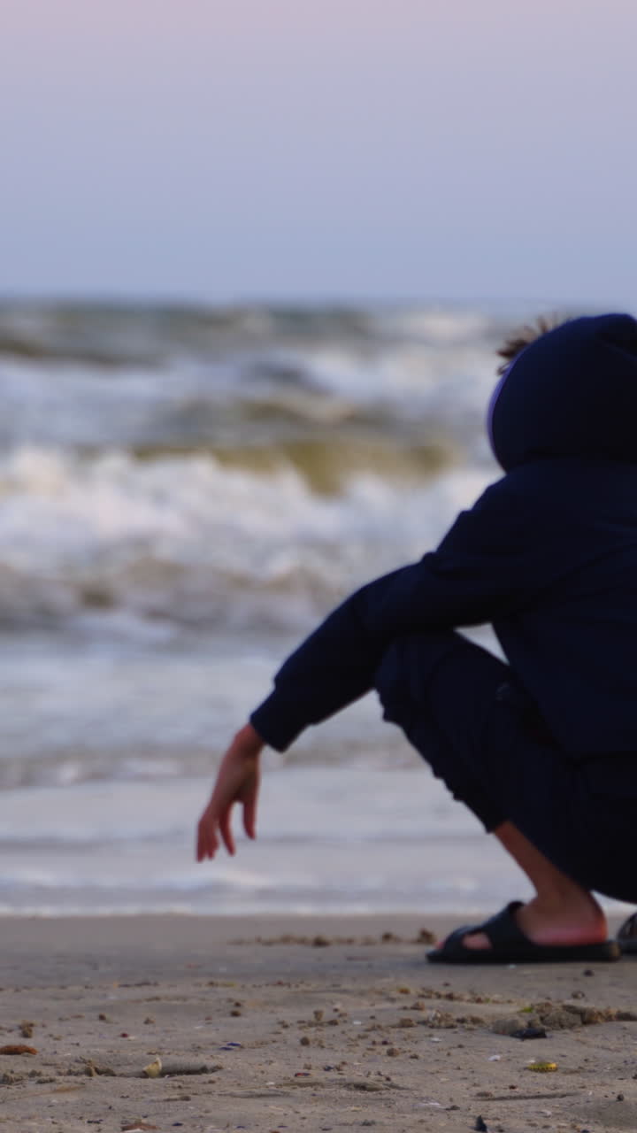 Boy resting near the sea at sunset. Child is painting on sandy beach on foamy waves background of the evening sea. Vertical video