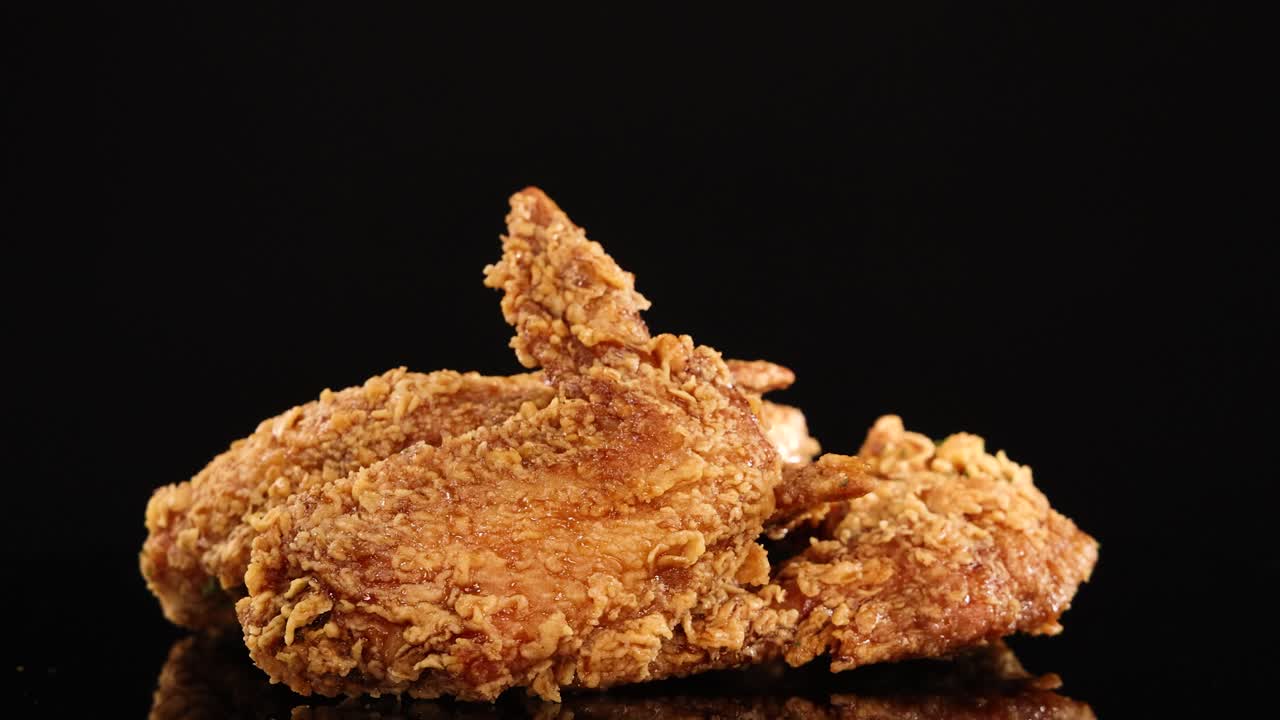 A hand reaches in and lifts a crispy fried chicken wing from a pile, shot in slow motion with dramatic studio lighting against a black background