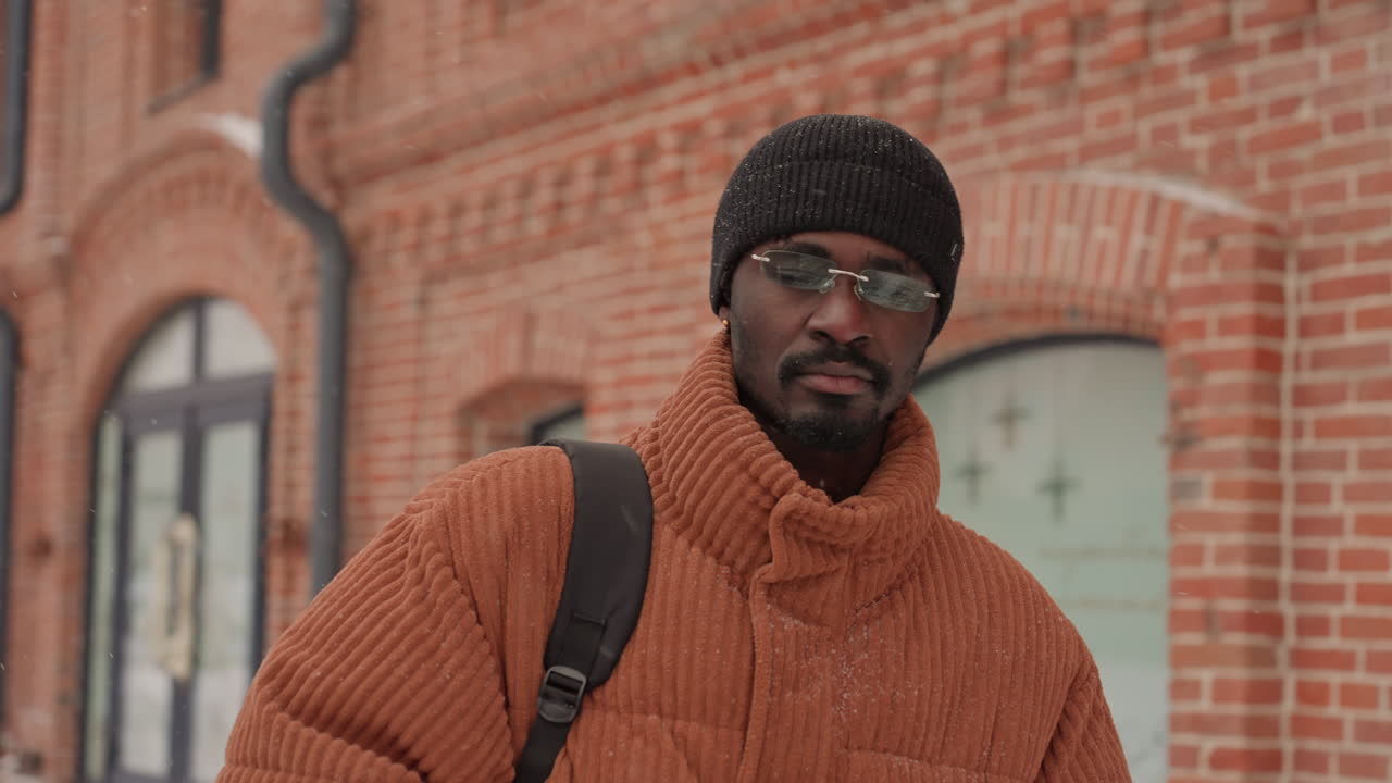 Uomo di colore robusto che cammina davanti a una vetrina di mattoni, cappotto oversize, zaino, berretto di maglia, occhiali a specchio, passo sicuro, respiro visibile nell'aria fredda, marciapiede urbano, pedoni sfocati, abbigliamento streetwear
