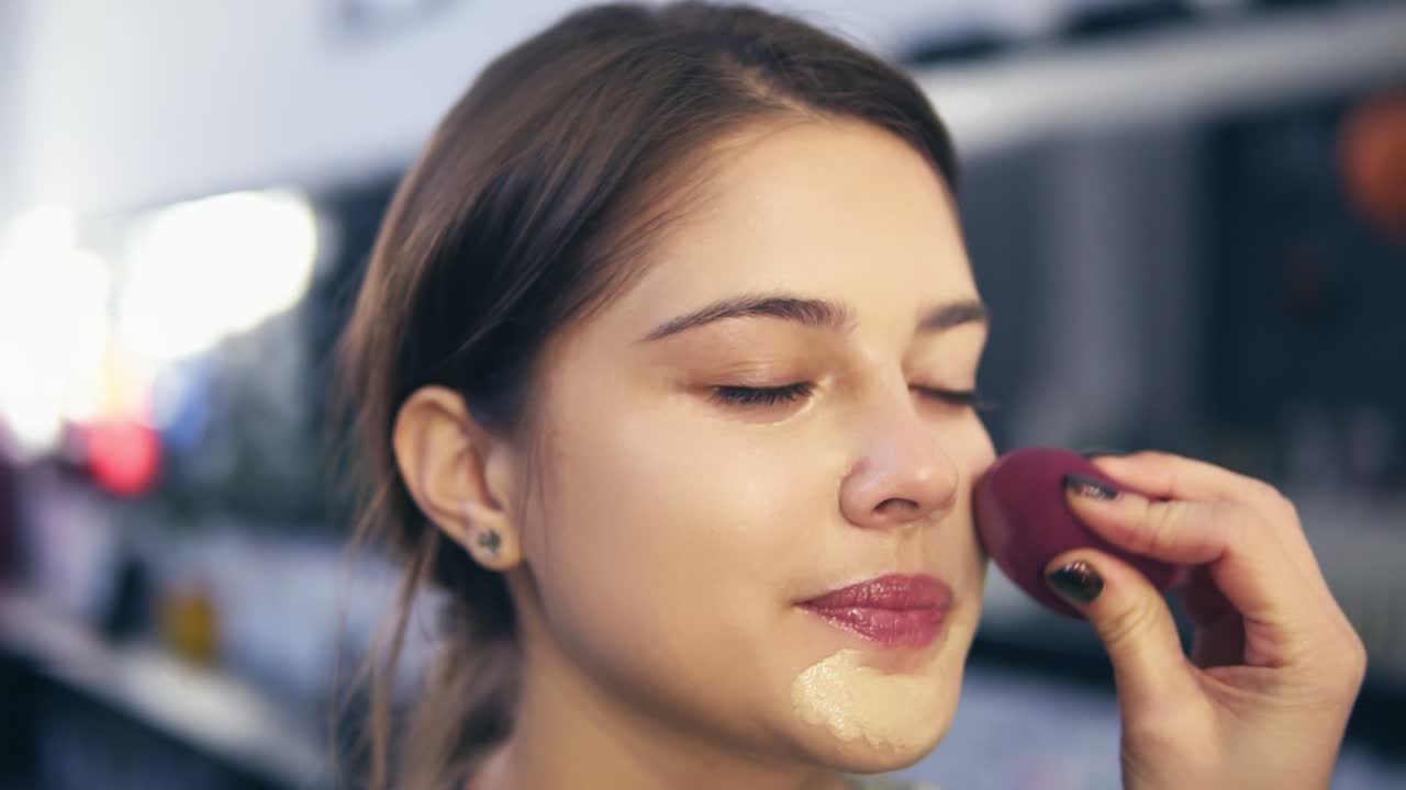 Unrecognizable visagist applying fluid foundation on young woman's face using sponge. Professional makeup in salon. Slow Motion