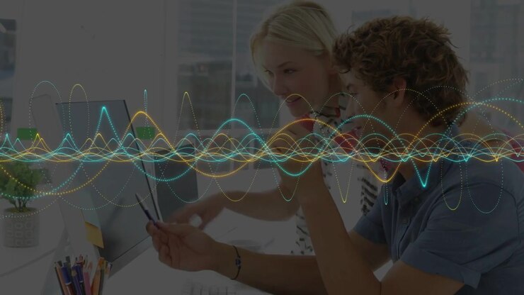 Мужчина работает за столом, женщина подходит и указывает на телефон и монитор, волновой график стимулирует бизнес-обзор.
