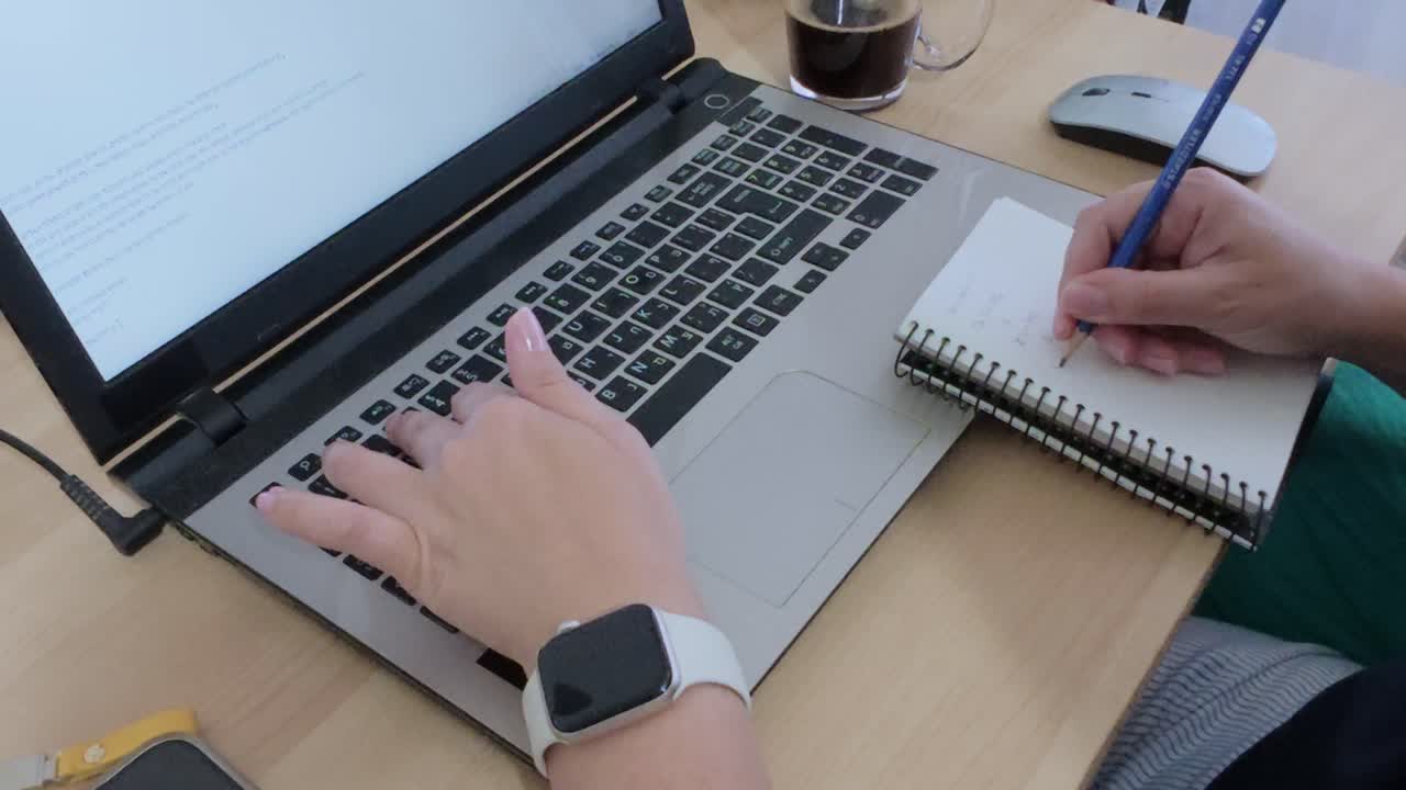 A modern desk setup with hands writing in a notebook, laptop, smartphone, and smartwatch