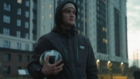 athlete holding soccer ball takes deep breath during urban training session, standing confidently in modern sport arena surrounded by tall residential buildings and dramatic evening sky