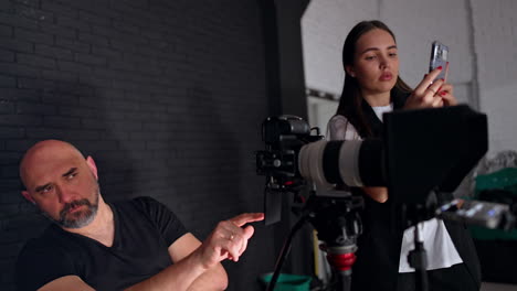caucasian bearded photographer sits looking at camera display. female assistant taking video on her smartphone.
