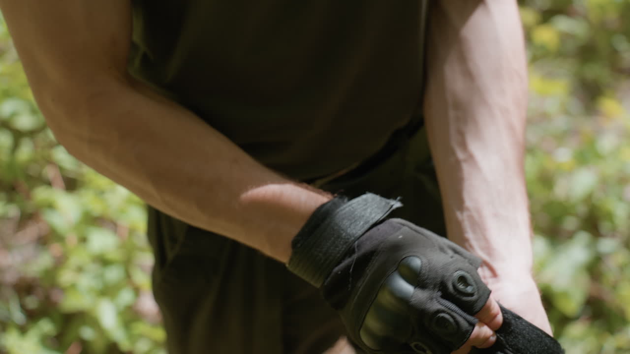 Young man outdoors adjusts tactical gloves and secures straps in forest environment, close up of hands showing careful precise grip, intent focus, skill ful readiness and strength for mission prep