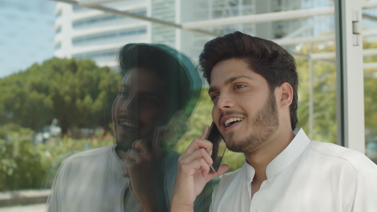 joven musulmán apoyado en una pared reflectante, hablando por teléfono.