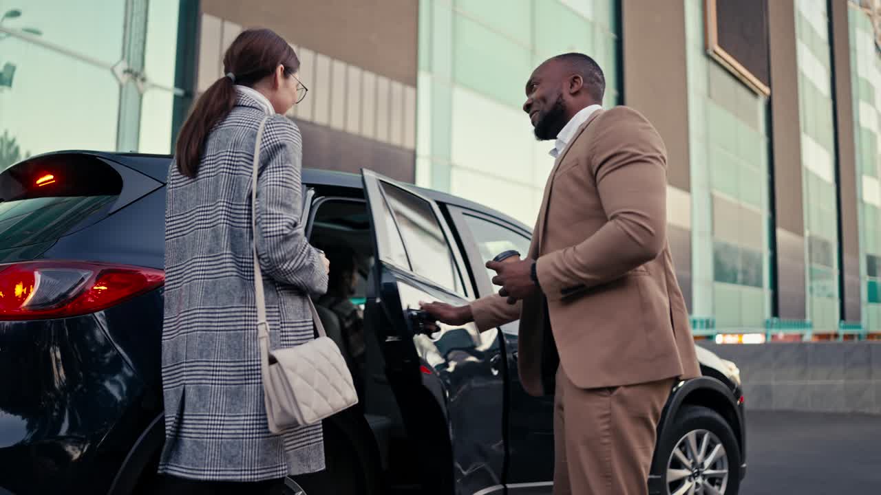 A man with black skin in a brown suit invites a girl to sit in the back seat of a modern Black car and sits down with her during their business trip around the city