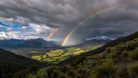 panoramic view of a vibrant rainbow over a lush green valley and snow-capped mountains under a dramatic sky