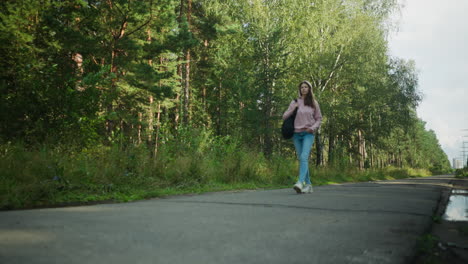 girl in walking calmly along quiet road near forest, holding black bag over shoulder with one hand in pocket, surrounded by greenery, trees, distant power lines, and puddles from recent rain