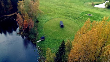 aerial backward moving shot over rectangular and cylindrical vacation cabin beside a pristine lake surrounded by colorful trees in rural countryside