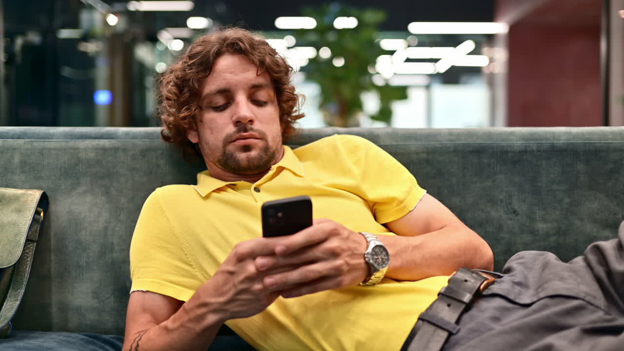 Mann i gul skjorte ligger på en sofa og scroller på telefonen sin