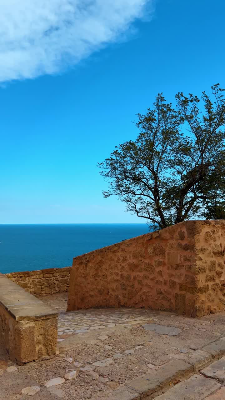 Terrazza in pietra con vista sul profondo blu del Mar Mediterraneo e cielo sereno. Vista del mare attraverso muri in pietra e rami di alberi verdi sotto un cielo azzurro.