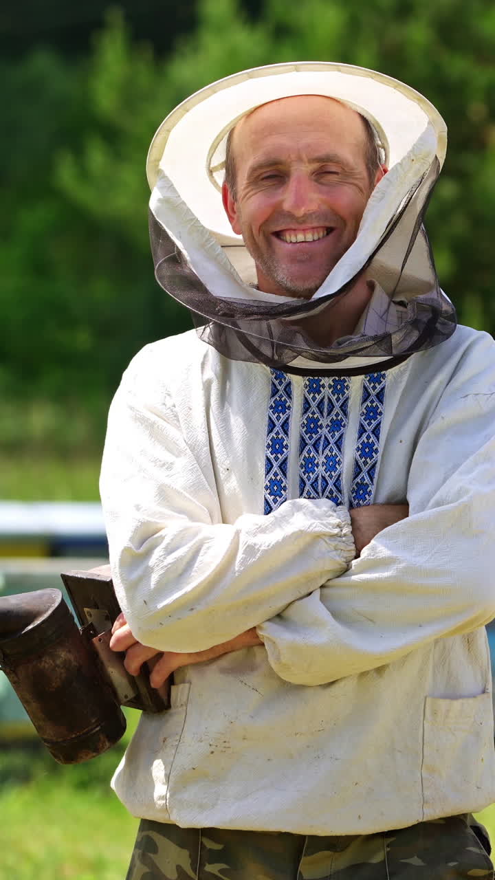 Beekeeper on apiary. Portrait of smiling man on outdoor hives background. Farmer apiarist in protective hat and in embroidery shirt looking on camera in summer day. Vertical video