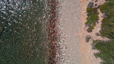 Vista-De-Arriba-Hacia-Abajo-De-Las-Transparentes-Olas-Del-Mar-Rompiendo-En-La-Playa-De-Guijarros-En-Cerdeña,-Italia---Toma-Aérea-De-Drones