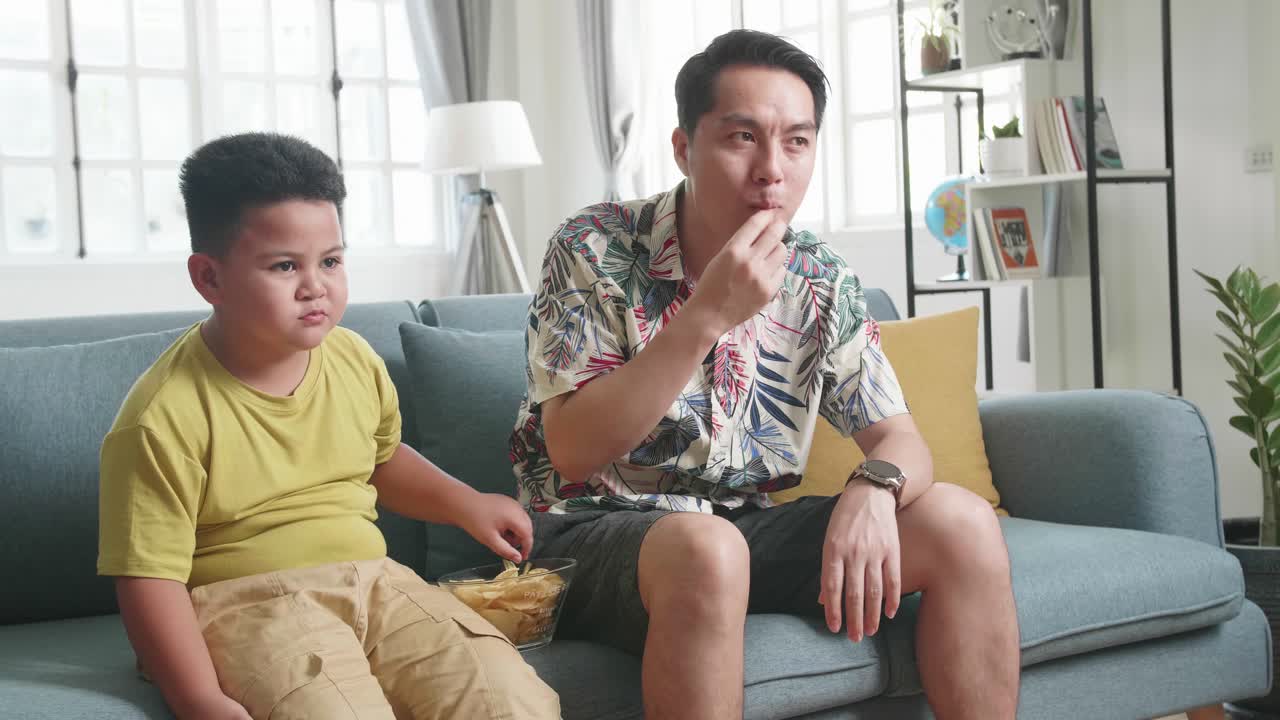 Happy Asian Father And His Son Are Watching Tv While Eating Potato Chips At Home