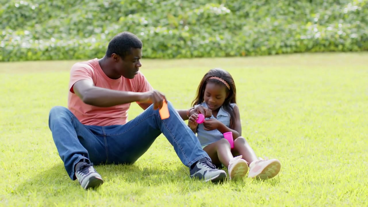 Blowing father and daughter playing with bubble wands on grassy lawn, with orange and pink bottles