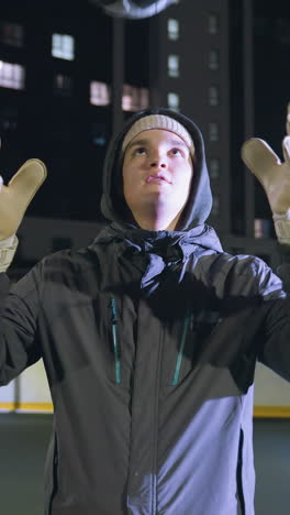 hombre con guantes lanza una pelota de fútbol al aire y la atrapa durante una sesión de entrenamiento nocturna en un campo urbano, el fondo iluminado