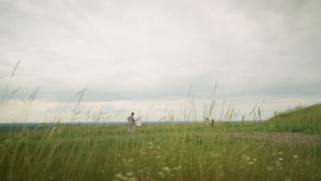 a craftsperson wearing a hat and checkered shirt is absorbed in painting on an easel in a peaceful meadow of tall grass under a cloudy sky. a woman in a hat and white gown sits calmly in a chair