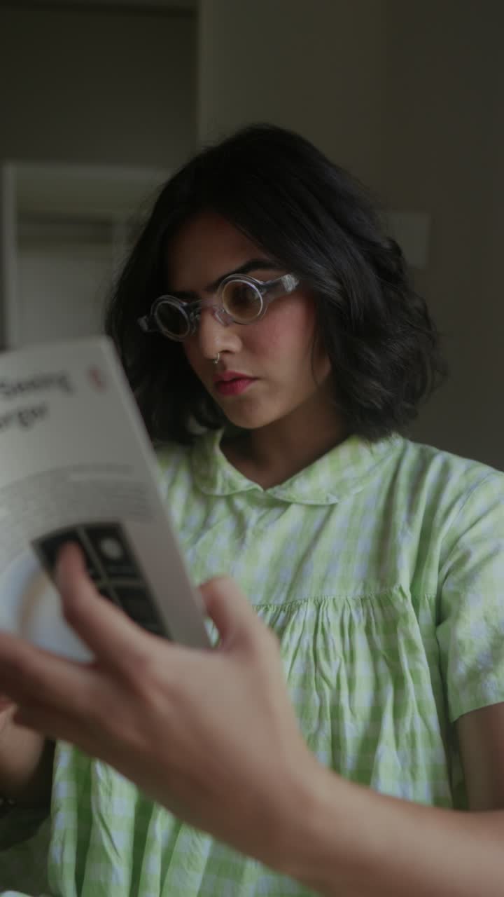 South Asian woman with short hair and clear glasses reading a booklet inside a softly lit room