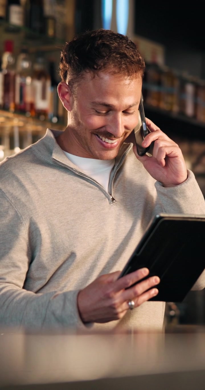 Young Man Talking on Smartphone While Looking at Tablet in Bar