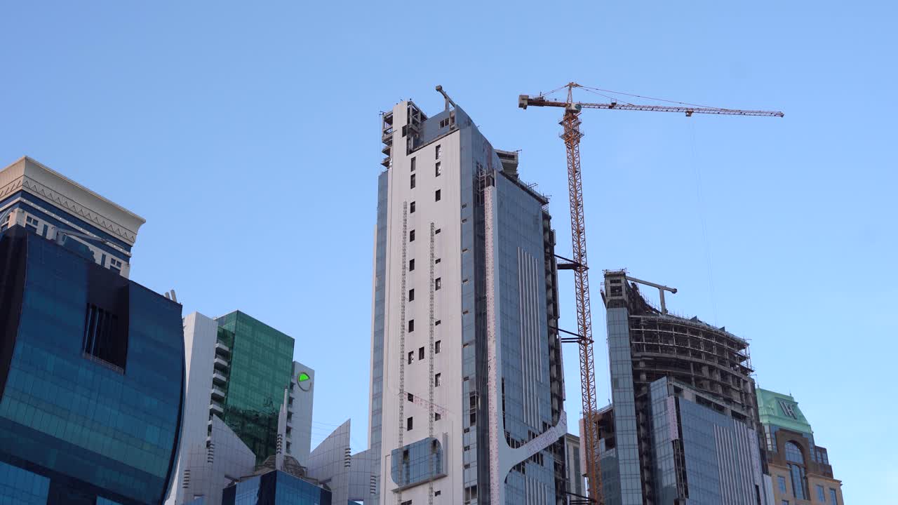 Una gru gialla alta lavora su un grattacielo in costruzione tra grattacieli moderni in vetro già completati nel centro di Doha, Qatar.