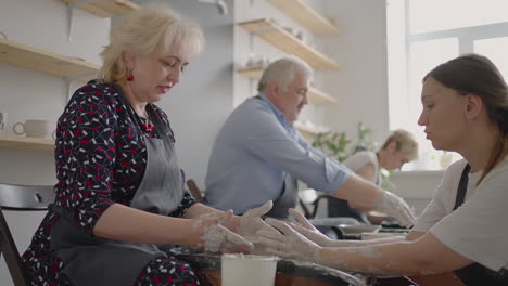 Medium-shot-of-middle-aged-ceramic-artist-teaching-group-elderly-Caucasian-woman-and-senior-man-how-to-wedge-clay-sitting-at-desk-in-art-studio.-People-enjoying-talking-at-work