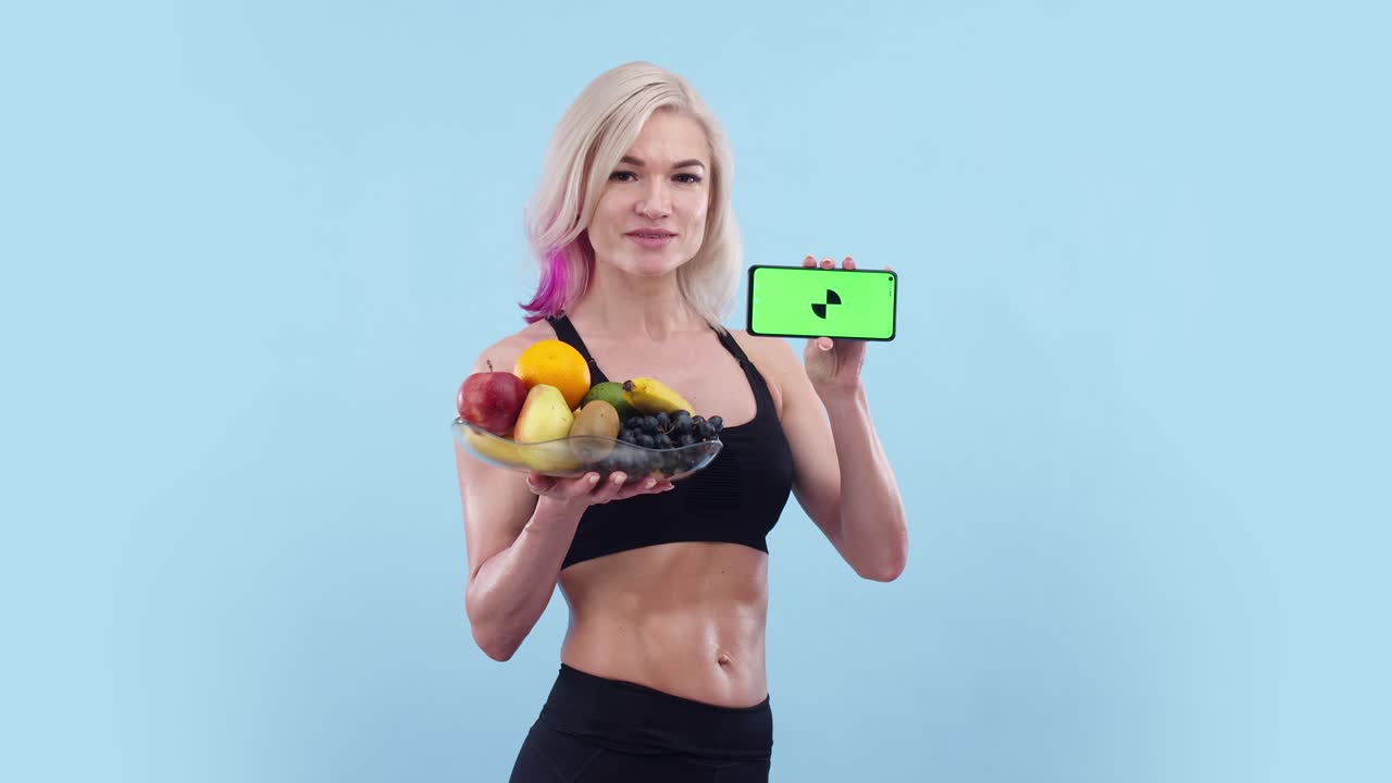 아름다운 운동선수 여성은 손에 과일 접시를 들고 초록색 화면 앱으로 전화를 보여줍니다. 금발 운동선수는 파란색 배경에서 스튜디오에서 과일과 전화를 들고 있습니다.