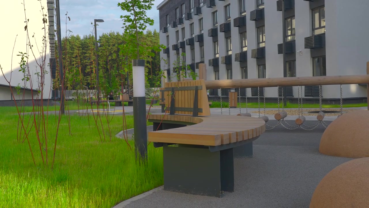 Un banc en bois se dresse au premier plan d'une cour d'immeuble paysagée avec de l'herbe fraîche, un jeune arbre, des garde-corps et une façade de bâtiment moderne dans une lumière douce.