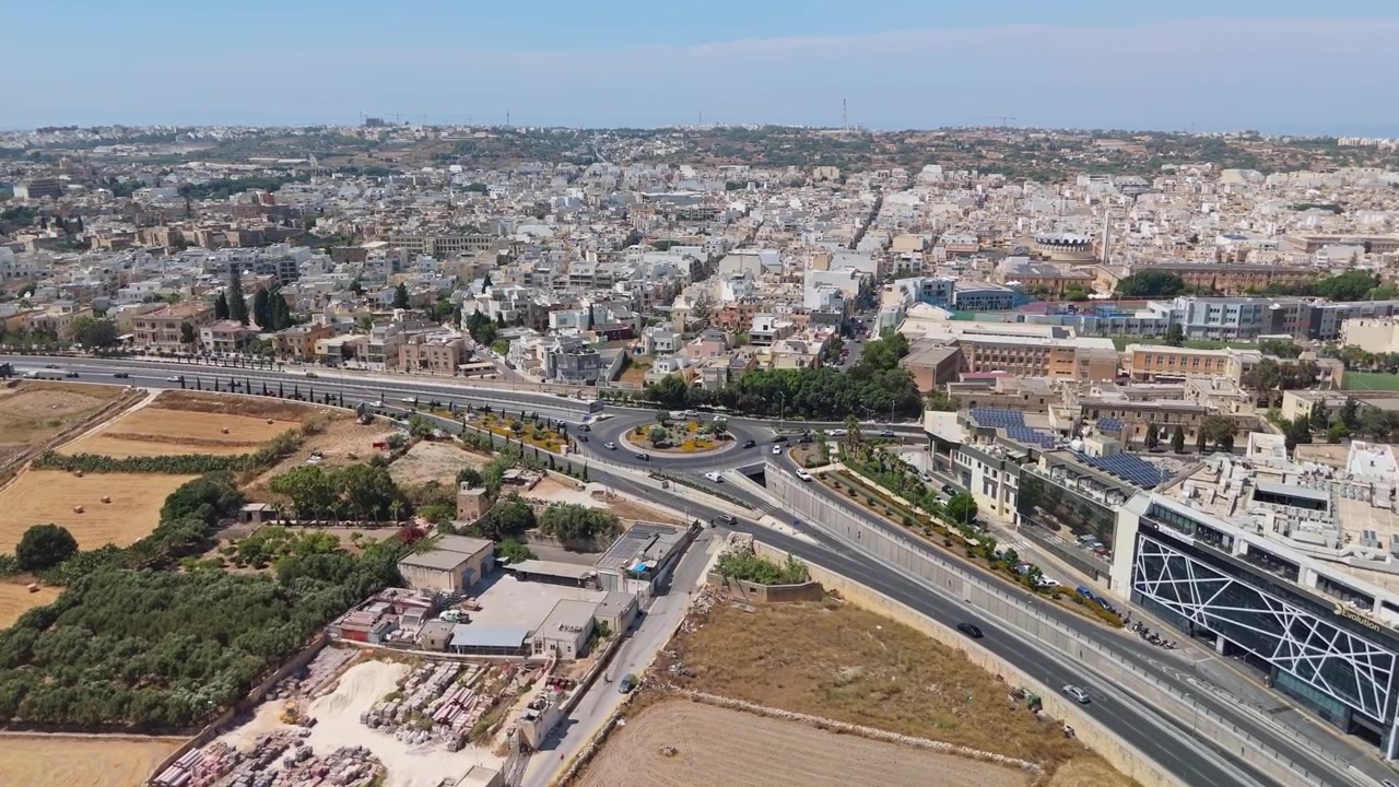 Rotatoria a Birkirkara e Balzan, Malta, con traffico sulle strade principali vicino a edifici commerciali e residenziali in un'area urbana mediterranea, ripresa con vista aerea