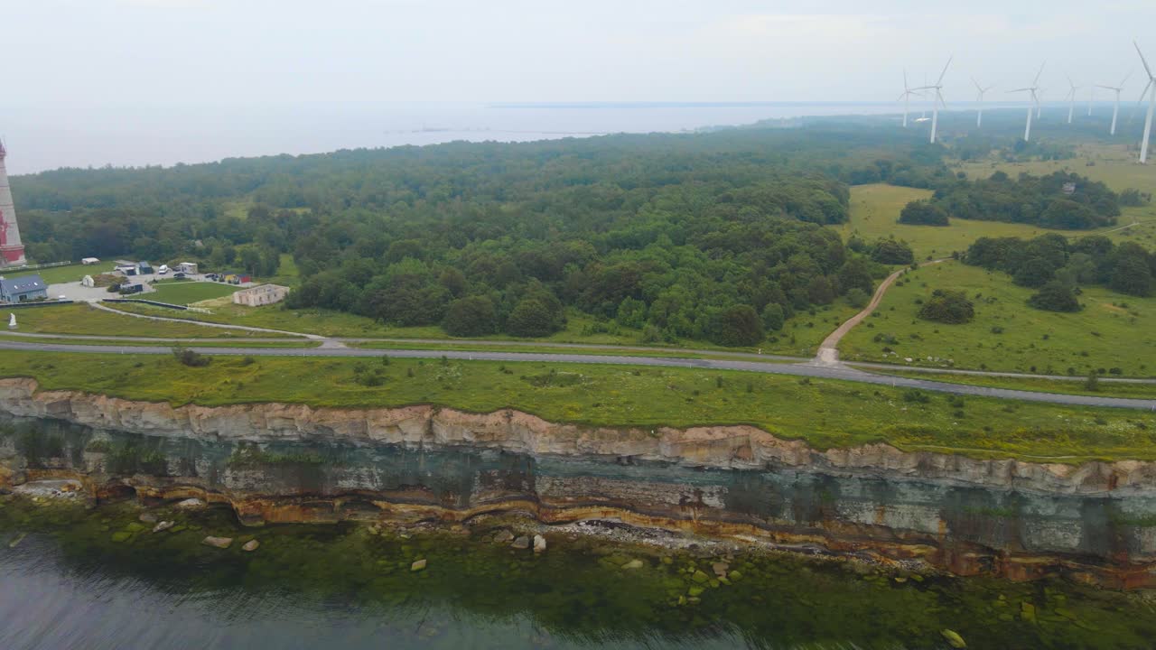 Aerial drone footage view over a steep cliff bank at Paldiski peninsula where renewable energy creating wind turbines are generating green energy and a historic red Pakri lighthouse is visible as well