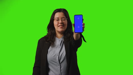 Smiling-Young-Businesswoman-Holding-Blue-Screen-Mobile-Phone-Towards-Camera-Standing-Against-Green-Screen-Background-1