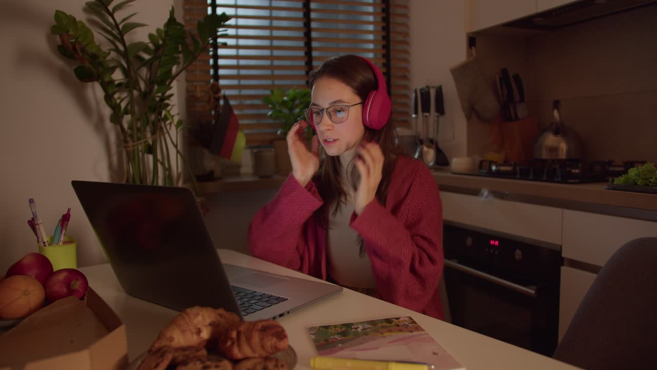 A confident brunette girl in red wireless headphones and glasses listens to the correct pronunciation of the native speaker and studies foreign languages online at home in the kitchen in front of a laptop near her there is a German flag
