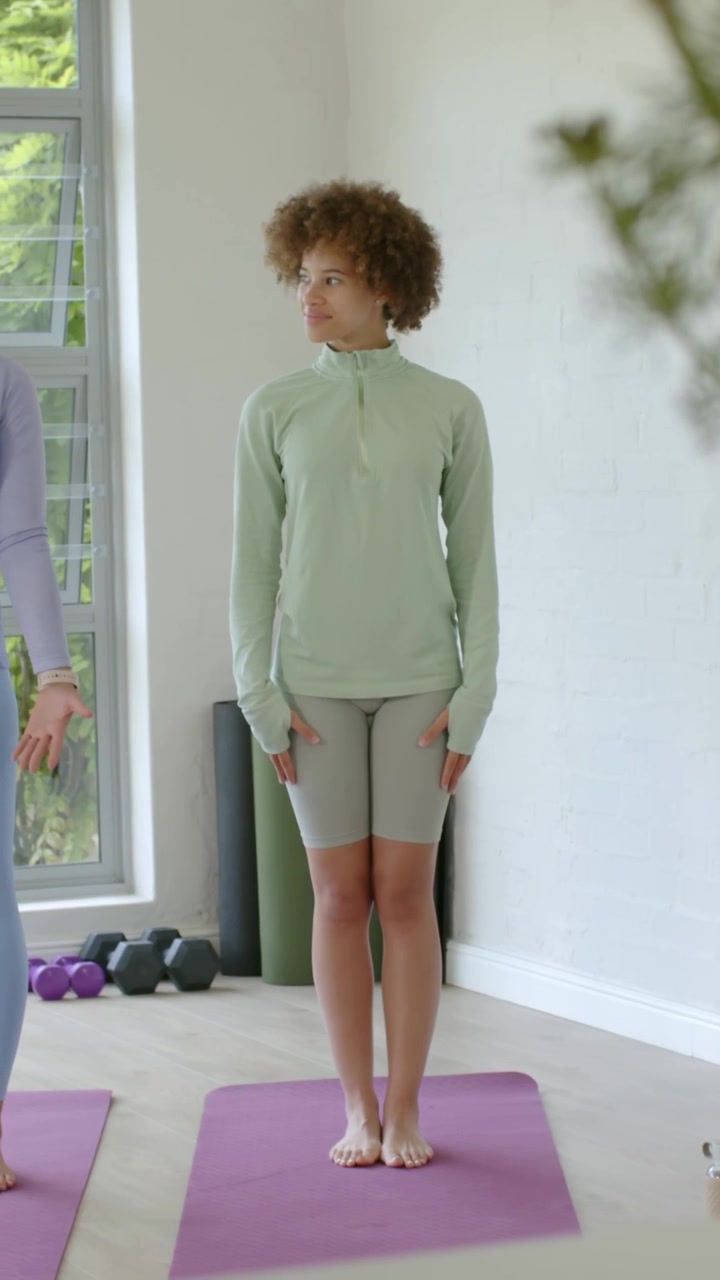Vertical video: Standing woman wearing green top on purple mat, following instructor gestures
