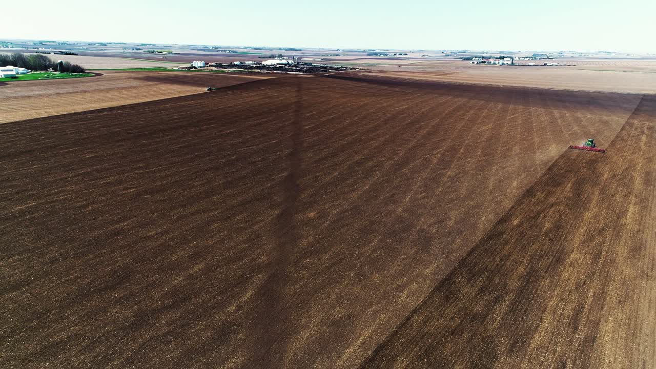 High drone shot overlooking a large freshly tilled field ready for planting with planter driving away in the distance