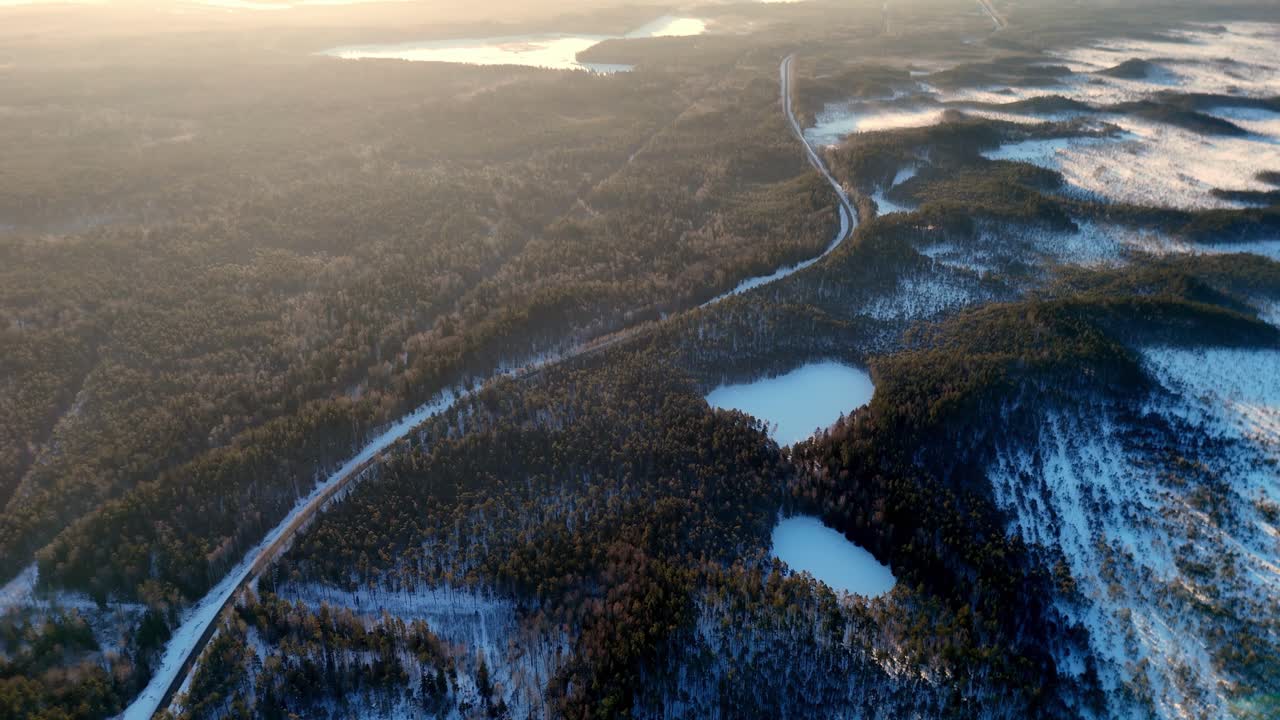 Luftfoto fra drone av en svingete vei gjennom en vidstrakt vinterskog med frosne innsjøer ved solnedgang fanger et fredelig snølandskap under mykt gyllent lys og tåkete atmosfære i en avsidesliggende nordlig villmark