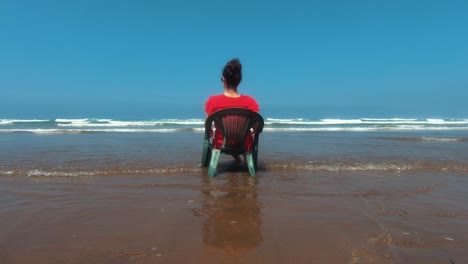 girl in read sitting on a green plastic chair on the beach of casablanca. filmed from behind.