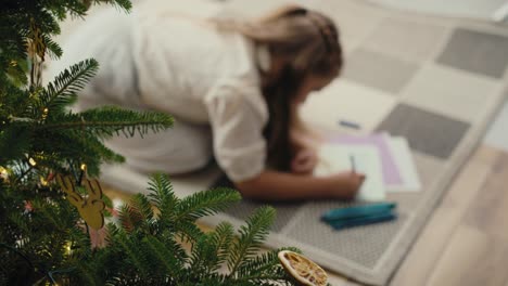 Top-view-of-little-caucasian-girl-lying-on-carpet-next-to-Christmas-tree-and-writing-a-letter-to-Santa-Claus.