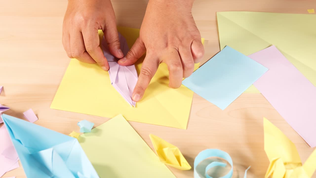 Person folds pastel origami paper, surrounded by colorful crafts, under bright, even lighting, overhead view
