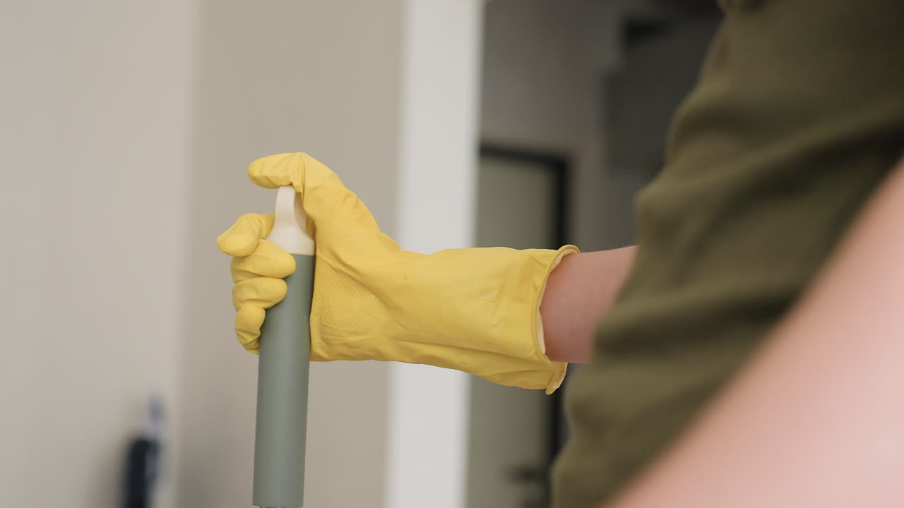 Partial view of cleaner wearing yellow glove holding mop handle with firm grip, blurred interior background visible, detail shot highlighting household cleaning activity