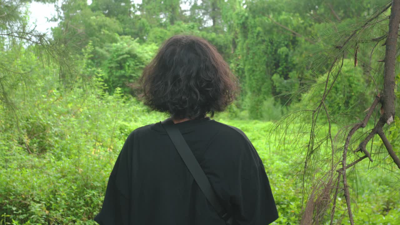 The camera moves forward toward a woman standing with her back to the lens, facing dense tropical greenery