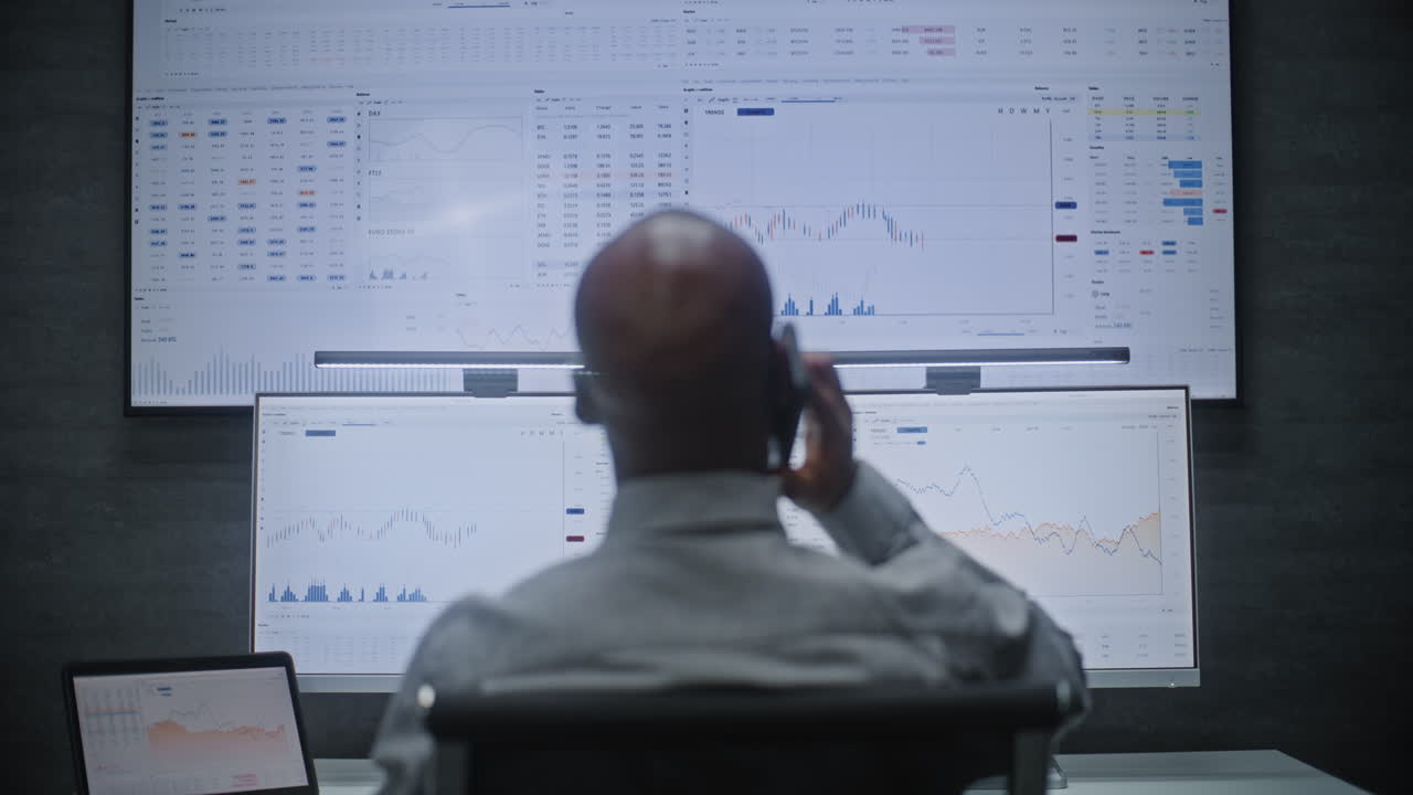 Stock Exchange Trader in Wireless Headphones Having Call, Monitoring Real-Time Stocks, Exchange Market Charts on Multi-Monitor Desk Setup. African American Investment Agent Working. Vertical Shot.
