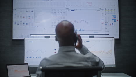 stock exchange trader in wireless headphones having call, monitoring real-time stocks, exchange market charts on multi-monitor desk setup. african american investment agent working. vertical shot.