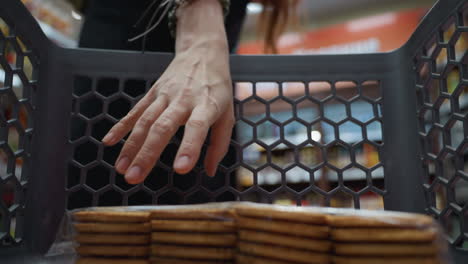 view of a lady in a black coat wearing a backpack, placing a pack of biscuits into a shopping trolley in a store. shot with a handheld camera