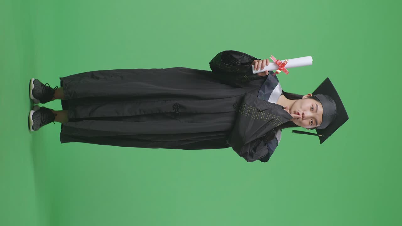 Studentessa asiatica laureata a figura intera in tocco e toga, con diploma e gesto 'shhh', su sfondo green screen in studio